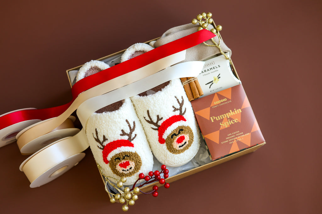 Holiday gift box featuring cozy reindeer-themed slippers, a pumpkin spice chocolate bar, vanilla caramels, cinnamon sticks, and festive decor with red and gold ribbon. Perfect for a warm and cheerful holiday gift